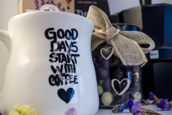 Caja regalo Musuka "Los buenos días empiezan con café" que contiene una cafetera Magefesa, una taza de cerámica artesanal en la que se lee "good days start with coffe", un paquete de galletas de cacao Lady Joseph, un paquete de Café Molido Bio “Tierra Madre Mujer”, de Oxfam Intermón Comercio Justo y un tarro de cacahuetes recubiertos de chocolate blanco y negro.