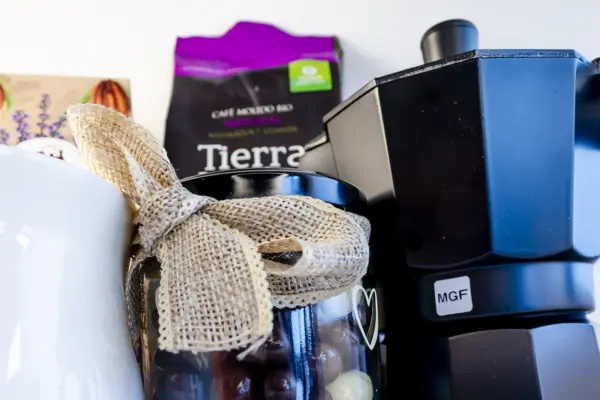 Caja regalo Musuka "Los buenos días empiezan con café" que contiene una cafetera Magefesa, una taza de cerámica artesanal en la que se lee "good days start with coffe", un paquete de galletas de cacao Lady Joseph, un paquete de Café Molido Bio “Tierra Madre Mujer”, de Oxfam Intermón Comercio Justo y un tarro de cacahuetes recubiertos de chocolate blanco y negro.