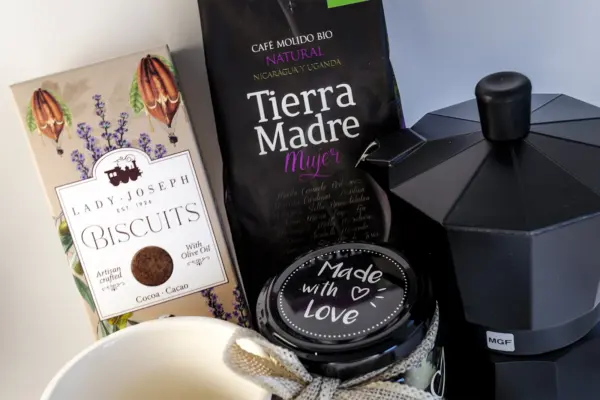 Caja regalo Musuka "Los buenos días empiezan con café" que contiene una cafetera Magefesa, una taza de cerámica artesanal en la que se lee "good days start with coffe", un paquete de galletas de cacao Lady Joseph, un paquete de Café Molido Bio “Tierra Madre Mujer”, de Oxfam Intermón Comercio Justo y un tarro de cacahuetes recubiertos de chocolate blanco y negro.