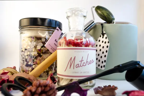 Caja de regalo Musuka "Mímate" que contiene: Una vela con olor a higuera silvestre y cedro, una herramienta para velas, un tarro de sales de baño con aroma a hibisco y rosa, un rodillo de masaje facial que se convierte en bolígrafo y una botella “Paddywax” con cerillas.