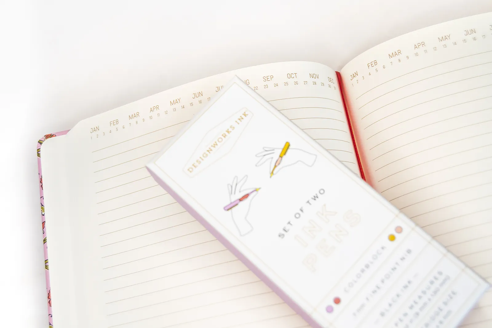 Agenda floral rosa abierta con estuche de bolígrafos Designworks Ink sobre sus páginas, una combinación de papelería bonita de la caja regalo Naroa.