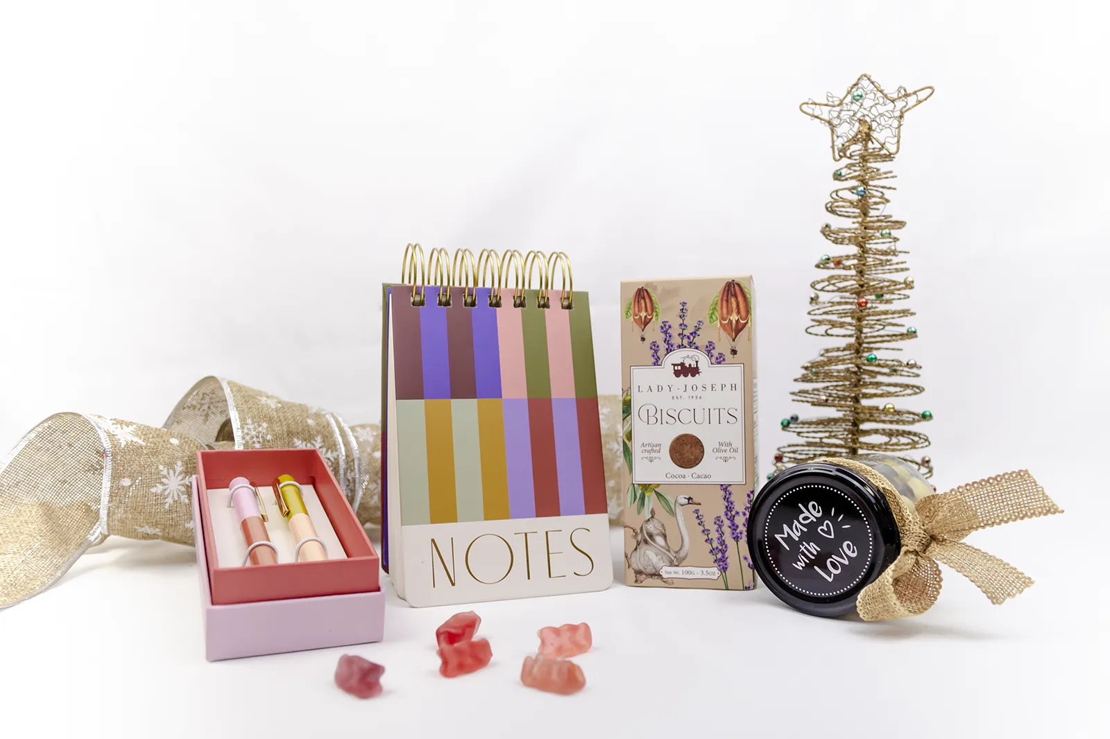 Caja Musuka “Regalo de Navidad 13" que contiene: Un bloc de notas con rayas de colores y anillas doradas. Un set de dos bolígrafos Designworks Ink en diferentes colores. Un tarro de cacahuetes recubiertos de chocolate blanco y negro. Un paquete de galletas de cacao Lady Joseph.