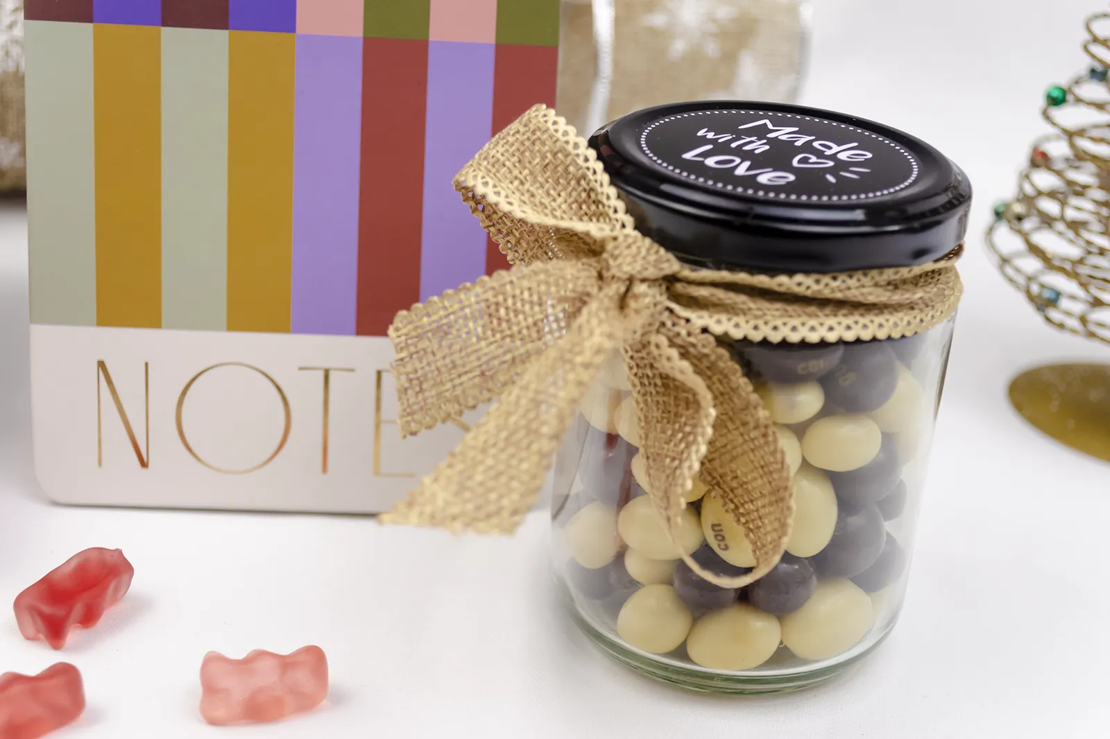 Caja Musuka “Regalo de Navidad 13" que contiene: Un bloc de notas con rayas de colores y anillas doradas. Un set de dos bolígrafos Designworks Ink en diferentes colores. Un tarro de cacahuetes recubiertos de chocolate blanco y negro. Un paquete de galletas de cacao Lady Joseph.