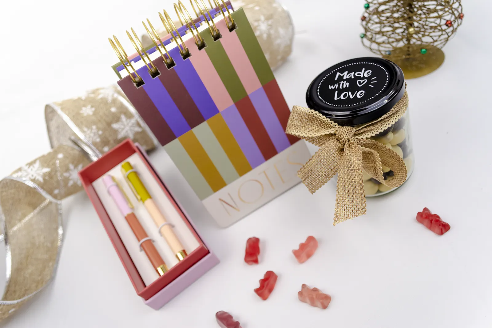 Caja Musuka “Regalo de Navidad 13" que contiene: Un bloc de notas con rayas de colores y anillas doradas. Un set de dos bolígrafos Designworks Ink en diferentes colores. Un tarro de cacahuetes recubiertos de chocolate blanco y negro. Un paquete de galletas de cacao Lady Joseph.