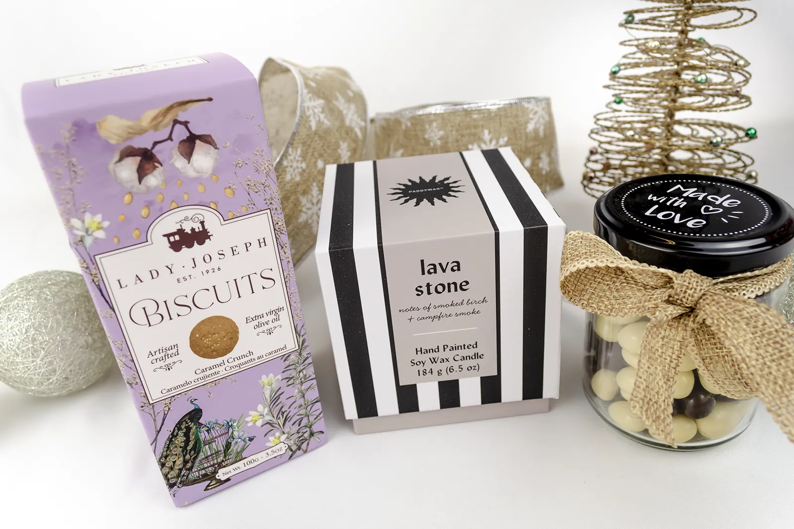 Caja Musuka “Regalo de Navidad 19" que contiene: Una vela Paddywax “Lava Stone”, en un vaso de rayas negras y blancas. Un paquete de galletas de caramelo crujiente Lady Joseph. Un tarro de cacahuetes recubiertos de chocolate blanco y negro.