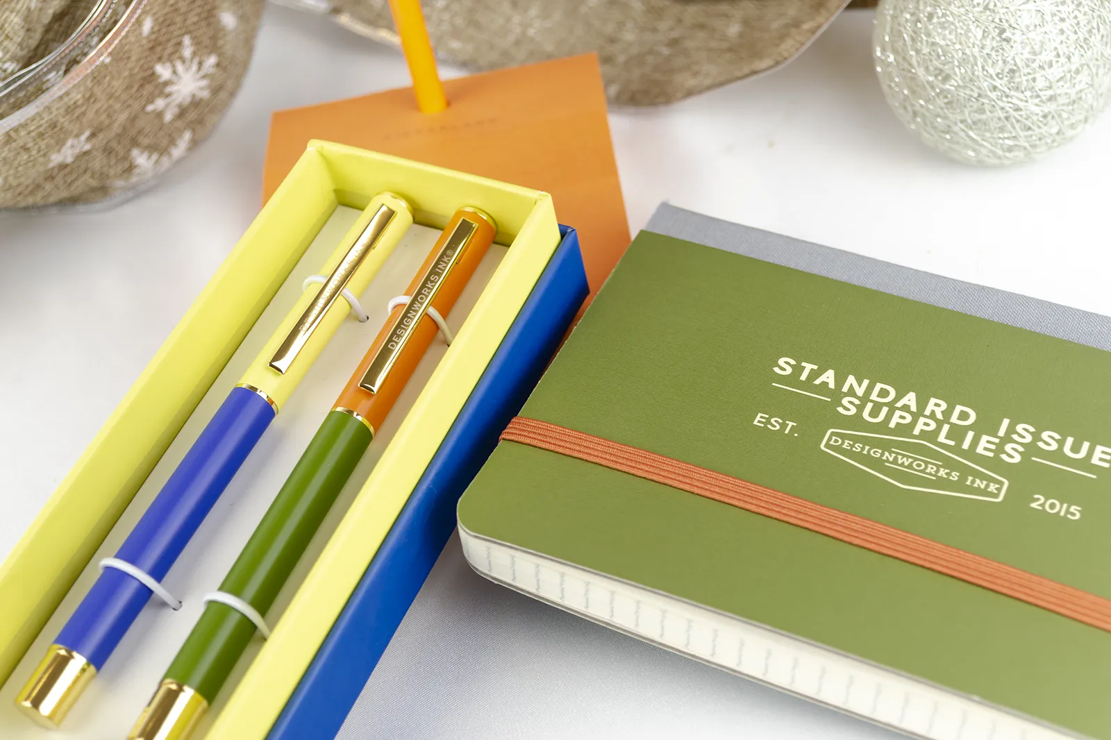 Caja Musuka “Regalo de Navidad 6" que contiene: set Kikkerland con un bloc de notas y un lápiz, con los que puedes montar una canasta. Un set de dos bolígrafos Designworks Ink, en tonos verdes y naranjas. Un cuaderno de bolsillo de tapa dura Designworks Ink con forma de pasaporte.