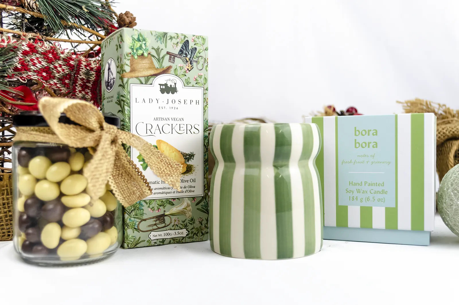 Caja Musuka “Regalo de Navidad 7" que contiene: crackers veganos Lady Joseph, vela Paddywax en un vaso de rayas verdes y blancas. Un tarro de cacahuetes recubiertos de chocolate blanco y negro.
