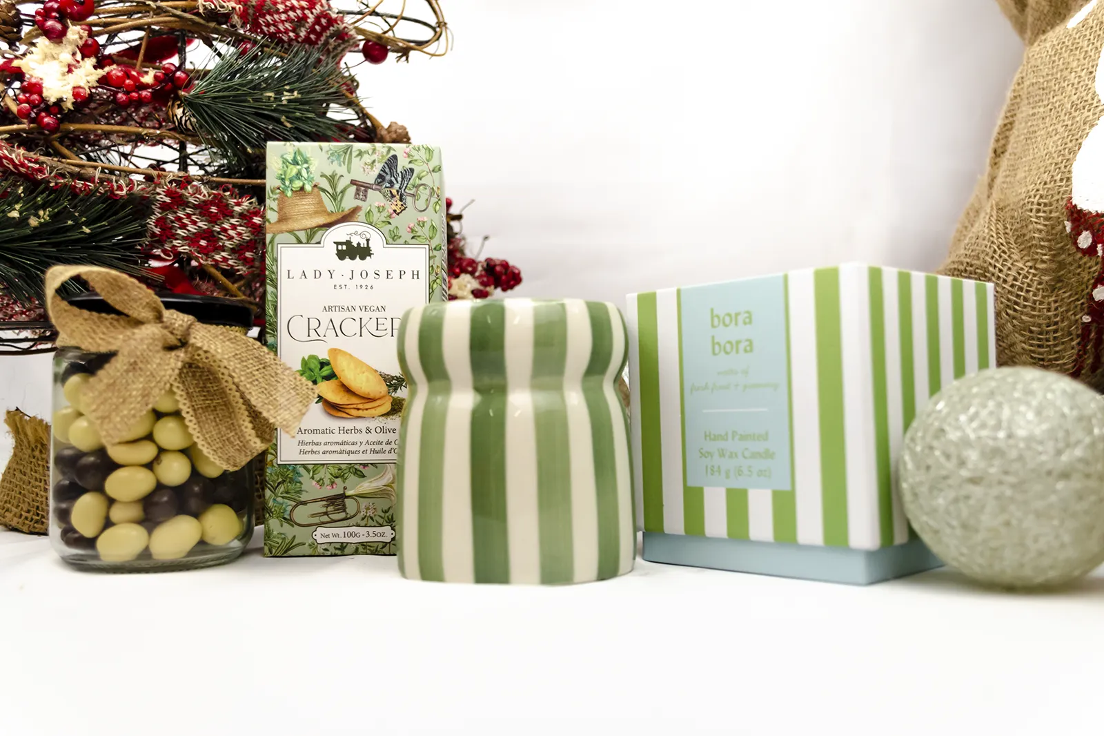 Caja Musuka “Regalo de Navidad 7" que contiene: crackers veganos Lady Joseph, vela Paddywax en un vaso de rayas verdes y blancas. Un tarro de cacahuetes recubiertos de chocolate blanco y negro.