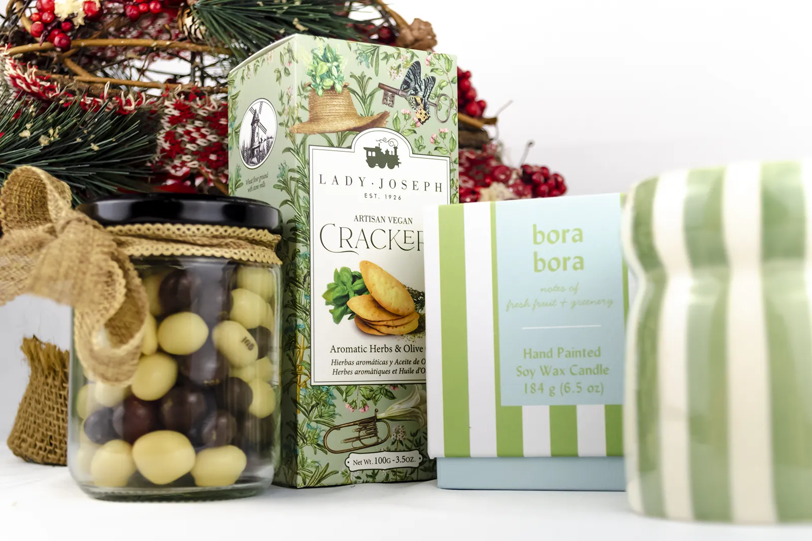 Caja Musuka “Regalo de Navidad 7" que contiene: crackers veganos Lady Joseph, vela Paddywax en un vaso de rayas verdes y blancas. Un tarro de cacahuetes recubiertos de chocolate blanco y negro.