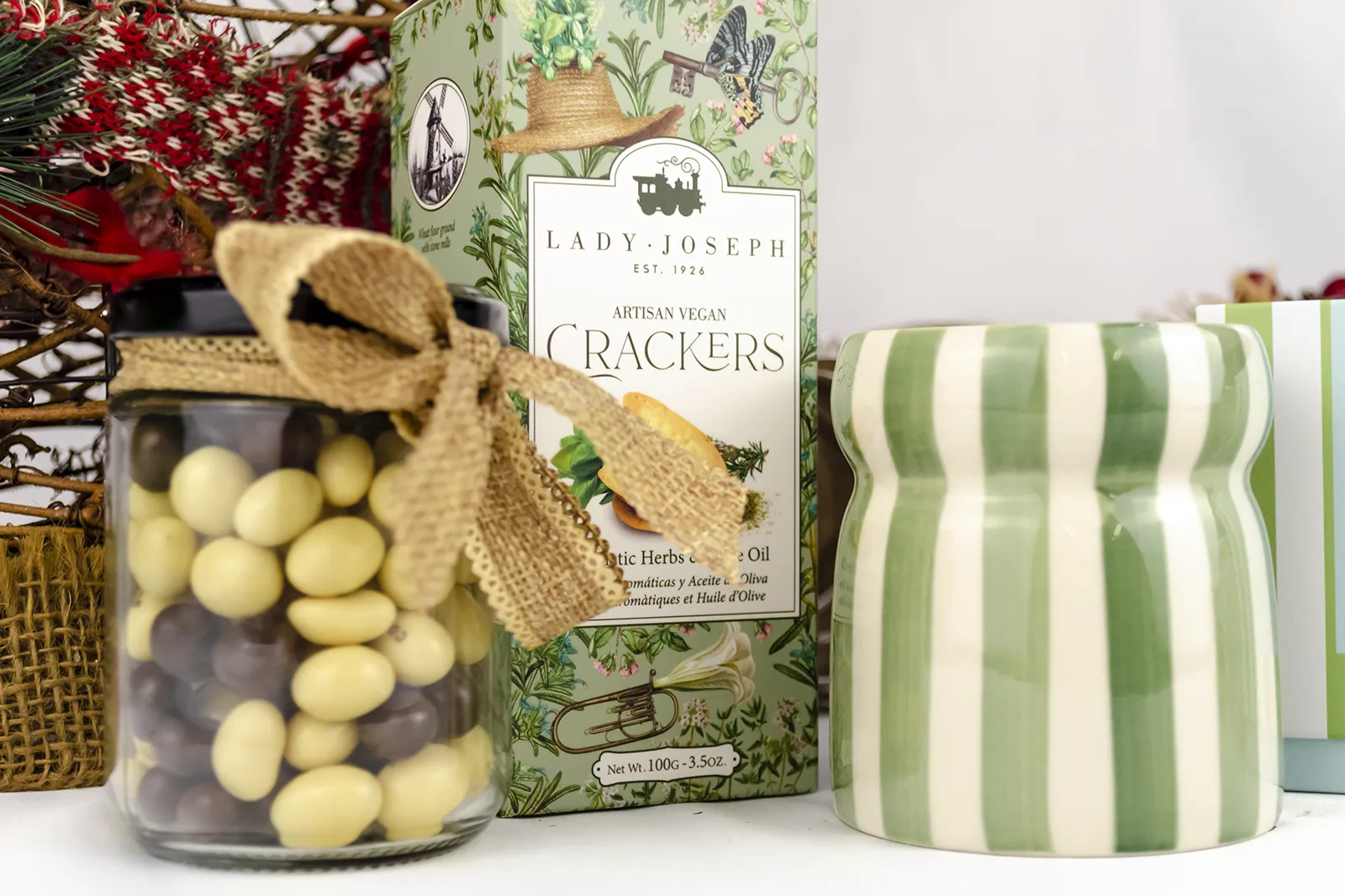 Caja Musuka “Regalo de Navidad 7" que contiene: crackers veganos Lady Joseph, vela Paddywax en un vaso de rayas verdes y blancas. Un tarro de cacahuetes recubiertos de chocolate blanco y negro.