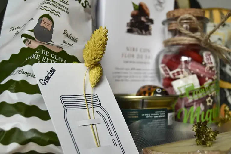 Regalo gourmet con galletas marineras, chocolate artesanal, paté de oca y botella personalizada junto a tarjeta de «Gracias» con flores preservadas