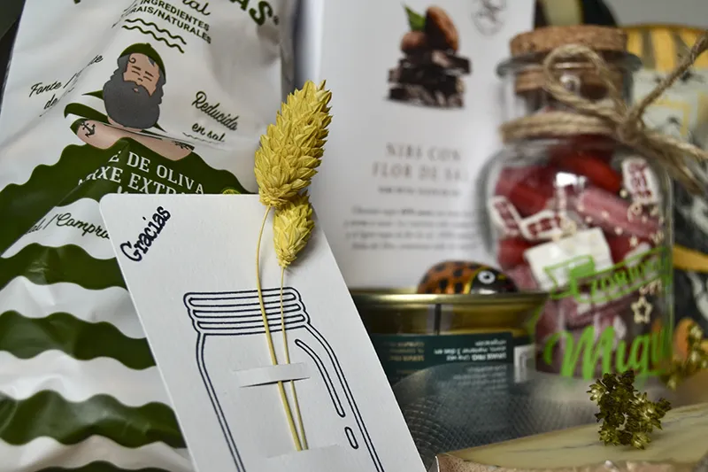 Regalo gourmet con galletas marineras, chocolate artesanal, paté de oca y botella personalizada junto a tarjeta de «Gracias» con flores preservadas